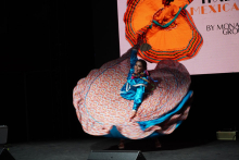 Osoba tańczy. Nosi niebieską bluzkę. Ma kolorową spódnicę z kwiatowym wzorem. Tło jest czarne. Twarz osoby jest zamazana.