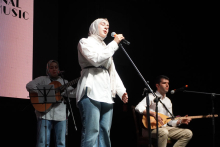 Trzy osoby stojące na scenie. Kobieta w środku nosi białą bluzkę i niebieskie dżinsy. Śpiewa do mikrofonu. Po lewej stronie osoba gra na gitarze klasycznej. Po prawej stronie osoba gra na instrumencie strunowym. Tło jest ciemne. 