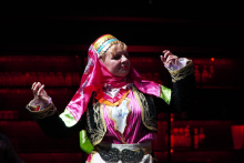 Kobieta tańcząca na scenie. Nosi tradycyjny, kolorowy strój ludowy. Ma chustę na głowie w kolorach różowym i żółtym. Górna część stroju jest różowa. Ma białe i złote zdobienia. Na rękawach są zielone akcenty. Osoba unosi ręce. Wygląda, jakby tańczyła lub wykonywała gesty. Tło jest ciemne. W tle widać czerwone światła.