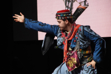 Mężczyzna w tradycyjnym stroju ludowym. Strój jest niebieski z czarnymi wzorami i czerwonymi akcentami. Mężczyzna na głowie ma czerwoną czapkę z kolorowymi ozdobami. Prawa ręka jest wyciągnięta do przodu. Lewa ręka spoczywa na biodrze. W tle widać duży ekran.