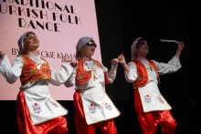 Trzy kobiety, które tańczą tradycyjny turecki taniec ludowy. Noszą białe koszule i czerwone spodnie. Mają białe fartuchy z kolorowymi haftami. Na głowach mają chustki. W tle widać duży ekran. Na ekranie jest napis: „TRADITIONAL TURKISH FOLK DANCE" 