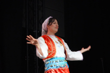 Kobieta na scenie. Ma na sobie tradycyjny strój ludowy. Nosi białą koszulę z długimi rękawami, czerwoną kamizelkę z haftem i niebieski pas ozdobiony złotymi monetami. Na głowie ma białą chustę. Tło jest ciemne. Po lewej stronie widać metalową konstrukcję sceny.