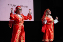Dwie kobiety ubrane w tradycyjne tureckie stroje taneczne. Stroje są czerwone i mają bogate złote zdobienia. Obie osoby noszą czerwone chusty na głowach. Ich ręce są uniesione do góry w geście tanecznym. W tle znajduje się ekran z napisem „BY TURECKIE KLIMATY”.
