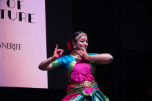 Młoda kobieta, która tańczy tradycyjny taniec. Ma na sobie kolorowy strój w odcieniach różu, zieleni i złota. Na głowie nosi ozdobę. Włosy są upięte w kok, ozdobiony czerwonym kwiatem. Tło jest ciemne. Po lewej stronie widać jasny ekran z napisem „OF TURE” i „ANERJEE”.