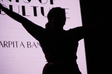 Sylwetka osoby z rozłożonymi rękami. Osoba stoi na tle jasnego ekranu. Na ekranie znajduje się tekst: „SPIRITS OF CULTURE”. Tło jest jasne, a postać widoczna jako ciemny kontur.