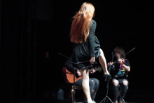 Kobieta z długimi, rudymi włosami tańcząca na środku sceny. Jest ubrana w czarną sukienkę. Ma podniesioną jedną nogę. Za nią siedzi dwie osoby. Jedna gra na gitarze akustycznej, druga na skrzypcach. Tło jest ciemne. Scena jest oświetlona jasnym światłem.