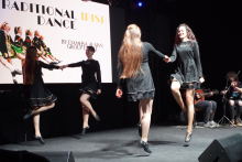 Cztery tancerki w czarnych sukienkach. Tańczą na scenie, trzymają się za ręce i unoszą nogi w powietrzu. W tle znajduje się duży ekran z napisem „Traditional Irish Dance”. Po prawej stronie sceny siedzi trzech muzyków. Grają na instrumentach.