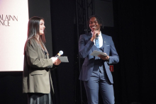 Młoda kobieta i młody mężczyzna stojący na scenie. Kobieta po lewej stronie ma długie, ciemne włosy. Nosi beżową marynarkę i trzyma mikrofon w prawej ręce. Mężczyzna po prawej stronie ma na sobie granatowy garnitur i krawat. Trzyma mikrofon w lewej ręce i kartki papieru w prawej. Tło jest ciemne. Po lewej stronie widać fragment ekranu.