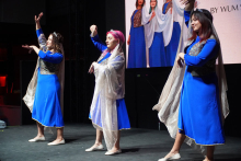 Trzy młode kobiety tańczące na scenie. Mają na sobie niebieskie sukienki z długimi rękawami. Na górze sukienek są złote wzory. Na ramionach mają przezroczyste, złote szale. Tancerki stoją w różnych pozach z uniesionymi rękami. Tło jest ciemne. Za nimi znajduje się duży ekran z obrazem innych osób w podobnych strojach.