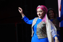 Młoda kobieta ubrana w niebieską suknię i kamizelkę z wzorem. Jedna rękę ma uniesioną do góry. Kobieta ma różowe włosy. 