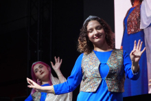 Dwie młode kobiety tańczące na scenie. Kobieta na pierwszym planie ma kręcone włosy. Nosi niebieską sukienkę i wzorzystą kamizelkę. W tle widać drugą osobę w podobnym stroju. 