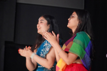 Dwie kobiety stojące obok siebie. Kobieta po lewej stronie nosi niebieską sukienkę z białymi wzorami i srebrną bransoletkę na nadgarstku. Kobieta po prawej stronie ma kolorową chustę w odcieniach tęczy i czerwoną spódnicę. Obie mają długie, ciemne włosy i klaszczą w dłonie. Tło jest ciemne, za nimi znajduje się czarna ściana lub zasłona.