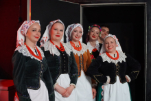 Sześć osób stojących w rzędzie. Mają na sobie tradycyjne stroje ludowe. Każda osoba nosi białą chustę z czerwonymi wzorami na głowie, białą bluzkę z koronkowym kołnierzem, ciemny gorset i biały fartuch. Dwie osoby mają zielone gorsety, jedna ma brązowy, a dwie czarne. Wszyscy mają czerwone korale na szyjach. Tło jest ciemne. Po lewej stronie widać fragment czerwonej ściany i jasnego przedmiotu.