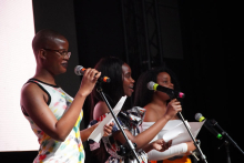 Trzy kobiety stojące na scenie. Każda trzyma mikrofon i kartkę papieru. Kobieta po lewej stronie ma krótkie włosy i nosi kolorową koszulkę bez rękawów. Kobieta w środku ma długie, ciemne włosy i sukienkę z wzorem. Kobieta po prawej stronie ma kręcone włosy. Również trzyma mikrofon i kartkę. Tło jest ciemne. Osoby są oświetlone światłem sceny.