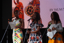 Trzy osoby stojące na scenie. Każda trzyma kartkę papieru i stoi przy mikrofonie. Kobieta po lewej stronie nosi kolorową sukienkę. Kobieta w środku ma sukienkę z wzorem w kółka i falbankami. Osoba po prawej stronie ma białą bluzkę z długim rękawem i żółtą spódnicę z niebieskimi wzorami. W tle znajduje się duży ekran. Na ekranie widać dwie osoby w tradycyjnych strojach afrykańskich i napis