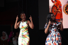 Dwie kobiety stojące na scenie. Kobieta po lewej stronie nosi obcisłą sukienkę w kolorowe wzory – białe, żółte, zielone i niebieskie. Kobieta po prawej stronie ma sukienkę z krótkimi rękawami w biało-niebiesko-czerwone wzory. Obie osoby mają uniesione ręce i wskazują palcami do góry. W tle znajduje się duży ekran z obrazem osoby w czerwonym stroju z czarnymi i złotymi elementami. 