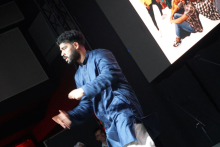 Mężczyzna w niebieskiej koszuli, który stoi na scenie. Jego twarz jest zamazana. W tle znajduje się duży ekran. Na ekranie wyświetlane jest zdjęcie grupy ludzi siedzących na podłodze. Scena jest ciemna. Światło pada na mężczyznę.