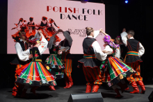 Grupa tancerzy wykonujących polski taniec ludowy. Mają na sobie tradycyjne stroje ludowe. Stroje składają się z kolorowych spódnic, kamizelek i czerwonych butów. W tle znajduje się duży ekran z napisem „POLISH FOLK DANCE”.