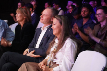 Grupa ludzi siedzących na białych krzesłach. Na pierwszym planie siedzi mężczyzna w ciemnym garniturze i kobieta w białej koszuli z długimi włosami. W tle widać inne osoby, niektóre klaszczą. Tło jest ciemne. Oświetlenie sceniczne tworzy kolorowe refleksy.