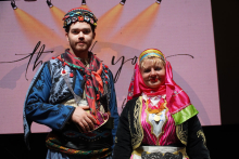 Po lewej stoi mężczyzna, po prawej kobieta. Są ubrani w tradycyjne stroje ludowe z Turcji. Mężczyzna ma niebieski strój z czerwonymi i zielonymi akcentami oraz kolorowy kapelusz. Kobieta ma na sobie różowy i czarny strój z ozdobami oraz różową chustę na głowie. Za nimi ekran projekcyjny z jasnoróżowym tłem. W tle widnieje napis „thank you”. Nad osobami świecą reflektory. 