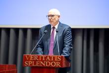 Rektor WUM. Mężczyzna W średnim wieku i okularach. Ma na sobie ciemny garnitur, białą koszulę i czerwonawy krawat. Stoi za mównicą i przemawiam. Mównica jest drewniana i ma napis "VERITATIS SPLENDOR" w złotym kolorze. W tle jest niebieski ekran multimedialny wiszący na szarych zasłonach.