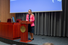 Kobieta w średnim wieku stojąca za mównicą. Kobieta ma na sobie różową marynarkę, czarną spódnicę i czarne buty. Mównica jest wykonana z ciemnego drewna i ma napis "VERITATIS SPLENDOR" oraz złoty emblemat na przodzie. W tle widać niebieski ekran projekcyjny. Na ekranie widoczne są białe elementy graficzne w prawym górnym rogu. Po lewej stronie mównicy znajduje się komputer stacjonarny z monitorem ustawionym bokiem do widza.