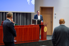 Trzech mężczyzn na sali konferencyjnej. Dwóch z nich jest odwróconych tyłem do obiektywu, trzeci stoi za mównicą i przemawia. Wszyscy są formalnie ubrani. Za mównicą znajduje się ekran multimedialny z niebieskim tłem i białym logo w prawym górnym rogu. Ściany sali są jasne, a podłoga ciemna.