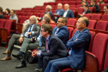 Grupa osób siedzących w czerwonych fotelach w audytorium. Większość osób jest ubrana formalnie, w garnitury i krawaty. Tło jest rozmyte, ale można zauważyć więcej osób siedzących dalej.