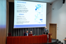 Sala konferencyjna z trzema mężczyznami na pierwszym planie. Dwóch siedzi za długim, drewnianym stołem, a jedna stoi za mównicą i prowadzi prezentację. Na ekranie projekcyjnym wyświetlony jest slajd z tekstem i mapami.