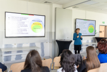 Kobieta w niebieskiej marynarce - prof. Hanna Szajewska - prowadzi prezentację. Stoi przed dwoma dużymi ekranami wyświetlającymi slajdy dotyczące sztucznej inteligencji, uczenia maszynowego i głębokiego uczenia. Publiczność składa się z kilku osób siedzących i zwróconych w stronę prezenterki. Na lewym ekranie widnieje tekst w języku polskim, a na prawym ekranie tekst w języku angielskim.