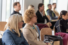 Grupa ludzi siedzących na krzesłach w jasnym pomieszczeniu podczas konferencji. Na pierwszym planie widoczne są dwie kobiety, jedna z blond włosami ubrana w niebieską kurtkę, druga - prof. Joanna Gotlib-Małkowska - z kręconymi włosami i naszyjnikiem w turkusowym kolorze. W tle znajdują się inne osoby.