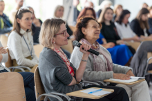 Grupa ludzi siedzi w sali konferencyjnej. Na pierwszym planie kobieta z blond włosami ubrana w szary sweter - prof. Dominika Nowis. Trzyma mikrofon. W tle widoczni są inni uczestnicy, siedzący i słuchający uważnie.