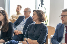 Grupa ludzi siedzi w sali konferencyjnej lub na spotkaniu. Osoby są ubrane w stroje biznesowe i zwrócone w stronę przodu - słuchają prelegenta. Na pierwszym planie kobieta w szarej sukience - dr Antonina Doroszewska. W tle ustawiony jest statyw z kamerą.