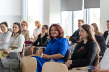 Grupa ludzi siedzących na krzesłach w jasnym pomieszczeniu podczas konferencji. W tle widać duże okna, przez które wpada naturalne światło. Na pierwszym planie znajduje się kobieta w niebieskiej sukience i okularach na głowie prof. Aneta Nitsch-Osuch. Pozostałe osoby są rozproszone po sali, niektóre z nich mają notatniki lub torby przy sobie."