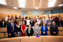 Grupowe zdjęcie około 35 osób w sali wykładowej. Część osób stoi, część siedzi. Uśmiechają się do zdjęcia. Większość osób jest ubrana formalnie: w garnitury, sukienki. Wiele osób ma na szyjach czerwone identyfikatory. W tle widoczne są drewniane ławki, jasne ściany i sufitowe oświetlenie.