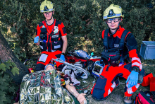 Dwóch ratowników medycznych w czerwonych uniformach i kaskach udziela pomocy osobie leżącej na ziemi, przykrytej folią termiczną. Poszkodowany jest podłączony do monitora EKG. Obok znajdują się torby medyczne, a w tle widoczne są krzewy.