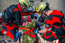 Dwóch ratowników medycznych w czerwono-czarnych uniformach i kaskach udziela pomocy osobie leżącej na bruku. Jeden wykonuje masaż serca, drugi obsługuje defibrylator. Poszkodowany jest przykryty folią termiczną i zabezpieczony pasami. W tle widoczny sprzęt ratunkowy.