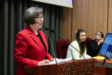 Kobieta stojącą przy mównicy w brązowym kolorze. Kobieta ubrana jest w czerwony żakiet. Na mównicy znajdują się dwa mikrofony. W tle, po prawej stronie, siedzą dwie osoby przy stole z komputerem. Na ekranie komputera widoczny jest fragment prezentacji.