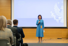 Woman in blue dress standing on stage during event. The woman holds a microphone in her right hand and a piece of paper in her left. Around her neck hangs a badge with an orange badge. Behind her is a large projection screen on which the logo of Warsaw Medical University is visible. The left side of the frame shows the backs of several people sitting in chairs, facing the stage. The floor is made of wood, and the walls are light brown in color.