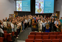 Grupa osób pozuje do zdjęcia w auli z czerwonymi siedzeniami. W tle dwa ekrany z napisem: ‘CRAIG MELLO RNAi: Ancient Mechanism, New Medicines’. 