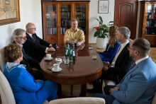 Siedem osób, władze rektorskie, dziekańskie i prof. Cameron C. Mello, w formalnych strojach siedzi przy owalnym stole w eleganckim biurze. Na stole filiżanki z kawą, butelki wody i szklanki. W tle drewniane regały z książkami i zamknięte drzwi.