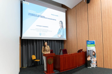 Sala konferencyjna z drewnianymi panelami na ścianach. Na środku znajduje się mównica, za którą stoi osoba w jasnym garniturze, przemawiająca do publiczności. Po prawej stronie mównicy znajduje się duży stół konferencyjny. Na ścianie za osobą przemawiającą wisi ekran projekcyjny.