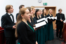 Grupa osób - członków chóru stojących w rzędzie wewnątrz auli. Mają otwarte usta, śpiewają. Wszyscy są ubrani w eleganckie, formalne stroje – garnitury, marynarki lub sukienki. Większość z nich trzyma otwarte czarne teczki lub segregatory, w których widoczne są białe kartki papieru.