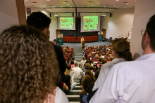 Wykład prof. Cameron C. Mello w auli. Publiczność siedzi na krzesłach i schodach, skierowana w stronę prelegenta. Przed nim dwa ekrany z obrazami przypominającymi komórki roślinne lub tkanki biologiczne.