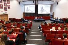 Duża sala wykładowa z czerwonymi siedzeniami, w której odbywa się symulacja rozprawy sądowej. Na przedzie sali znajdują się dwa duże ekrany wyświetlające obraz młotka sędziowskiego oraz tekst "Symulacja rozprawy". Kilka osób siedzi przy stołach z przodu sali, reprezentując różne role w symulacji. Publiczność składa się ze studentów.