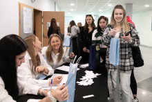 Po lewej stronie przy stole siedzą trzy osoby, mające przed sobą dokumenty, długopisy i białe kartki papieru. Po prawej stronie stoi kilka osób trzymających niebieskie torby i rozmawiających ze sobą. W tle widoczny jest korytarz, po którym poruszają się inne osoby w różnych kierunkach.