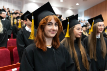 Grupa absolwentów w audytorium. Wszyscy ubrani są w czarne togi akademickie oraz birety z żółtymi chwostami. Na pierwszy planie po lewej jest młoda dziewczyna z rudymi włosami, która uśmiecha się do zdjęcia.