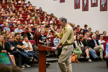 Mężczyzna, prof. Cameron C. Mello, w żółtej koszuli przemawia przy mównicy do licznej publiczności w audytorium. W tle rzędy czerwonych foteli zajętych przez słuchaczy oraz zdjęcia w ramach na ścianie.