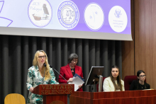 Cztery kobiety na sali konferencyjnej. Jedna stoi przy brązowej mównicy. Ma jasne włosy, nosi okulary, ubrana jest zielono-białą sukienkę. Przemawia do mikrofonu. Za nią widać trzy inne kobiety.W tle znajduje się ekran projekcyjny z czterema logotypami.