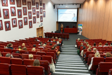 Sala wykładowa z rzędami czerwonych siedzeń, w większości zajętych przez ludzi. Na przedzie sali stoi prelegent przy mównicy, wygłaszający prezentację. Na ekranie za prelegentem wyświetlany jest slajd z tekstem i obrazem kobiety. Ściany są ozdobione oprawionymi w ramki portretami.